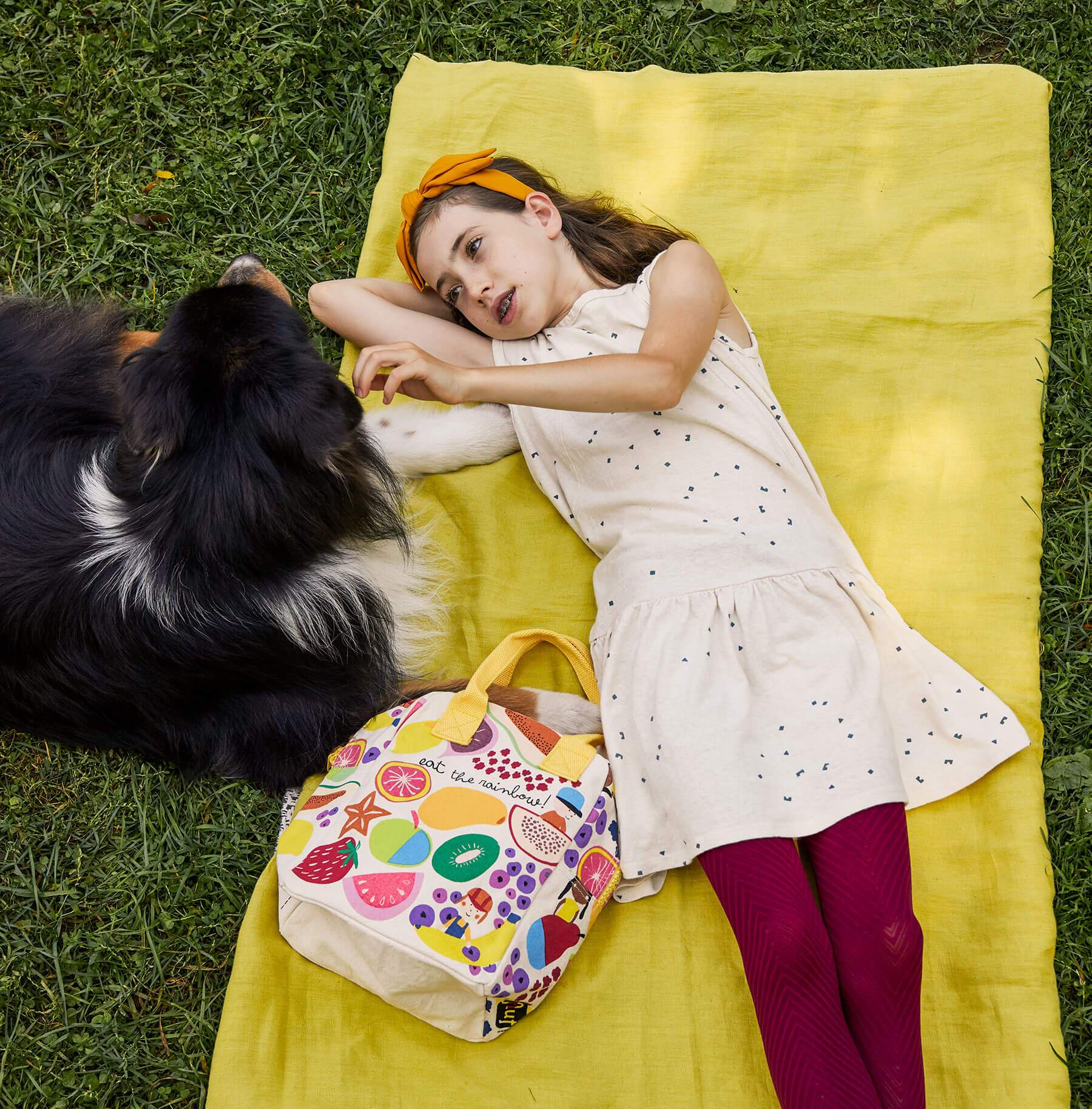 A girl on a yellow picnic blanket with Eat the Rainbow Lunch Bag playing with a dog