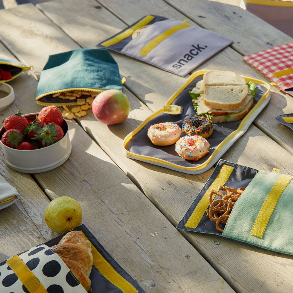 Fluf snack bags with various designs showcased on a table, filled with pretzels and croissants. One open bag reveals food-safe lining